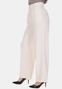 Pantaloni a gamba larga in tessuto color crema, con pieghe e vita alta, abbinati a scarpe nere con tacco alto. Tessuto liscio e vestibilità rilassata.