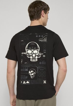 Mann trägt ein schwarzes T-Shirt mit weißem Grafikdesign von zwei Totenköpfen und abstrakten Tech-Stil-Elementen auf dem Rücken.