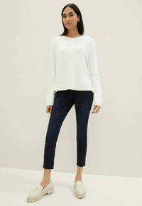 Žena oblečená v bílém svetru s dlouhým rukávem, tmavých slim-fit kalhotách a světlých mokasínech, stojící na jednobarevném pozadí.