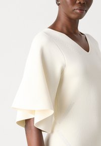 Cream-colored knit top with a V-neck and flared sleeves. Features subtle textured accents and a smooth finish.
