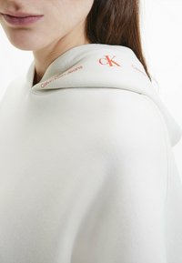 Sudadera gris claro con una textura suave, con el logotipo naranja "Calvin Klein Jeans" en el cuello y un distintivo logo cK.