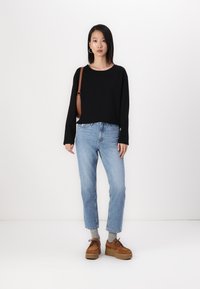 Svart langermet topp, lyseblå rette jeans og brune platåsko. Inkluderer en brun håndveske og grå sokker. Ensfarget bakgrunn.