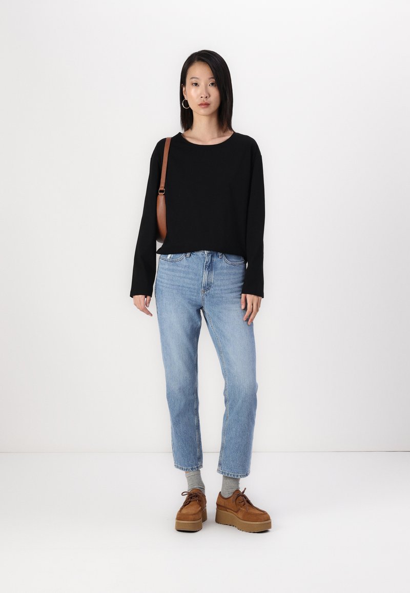 Svart langermet topp, lyseblå rette jeans og brune platåsko. Inkluderer en brun håndveske og grå sokker. Ensfarget bakgrunn.