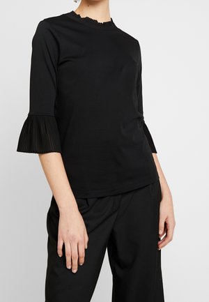 Woman wearing a black top with pleated 3/4-length sleeves and a ruffled neckline, paired with black pants against a plain background.