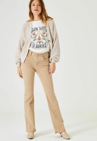Beige uitlopende jeans gecombineerd met een witte graphic t-shirt en een lichtbeige geribbelde cardigan, voorzien van grote knopen en textiele stof.