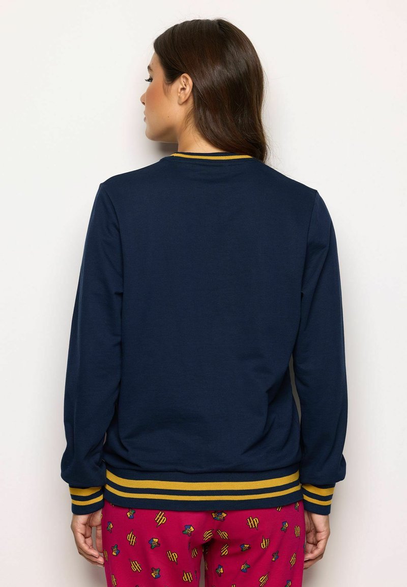 Vrouw met de rug naar de camera, gekleed in een navyblauwe trui met gele gestreepte manchetten en zoom, en rode broek met een patroon van "HP"-logo's en huisverleningen.