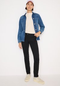 Giacca di jeans blu con chiusura a bottoni, abbinata a un maglione beige e jeans neri slim-fit, completata da sneakers di colore chiaro.