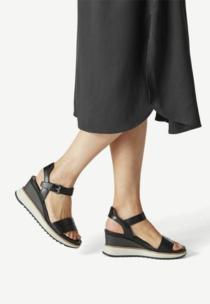 Damskie nogi w czarnych sandałach na koturnie z paskami na kostce, w stojącej pozycji, w czarnej spódnicy o średniej długości, na białym tle.