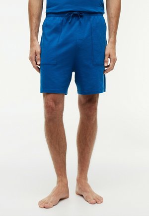 Hombre con pantalones cortos azules con cordón y bolsillos delanteros parche, de pie descalzo contra un fondo blanco.