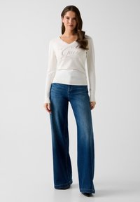 Weißer Pullover mit langen Ärmeln und „Guess“-Logo, kombiniert mit dunkelblauen Jeans mit weitem Bein. Beide Kleidungsstücke haben glatte Texturen und minimalistische Details.