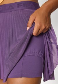 A hand holds up a purple athletic skirt, revealing an inner layer. The fabric features a textured waistband with the logo "FILA."