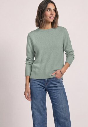 Femme aux cheveux bruns mi-longs portant un pull vert clair et un jean bleu, debout avec une main dans la poche sur un fond uni.