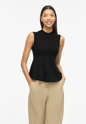 Femme souriante aux longs cheveux foncés portant un top noir sans manches à basque et un pantalon beige à jambes larges, debout les mains dans les poches.