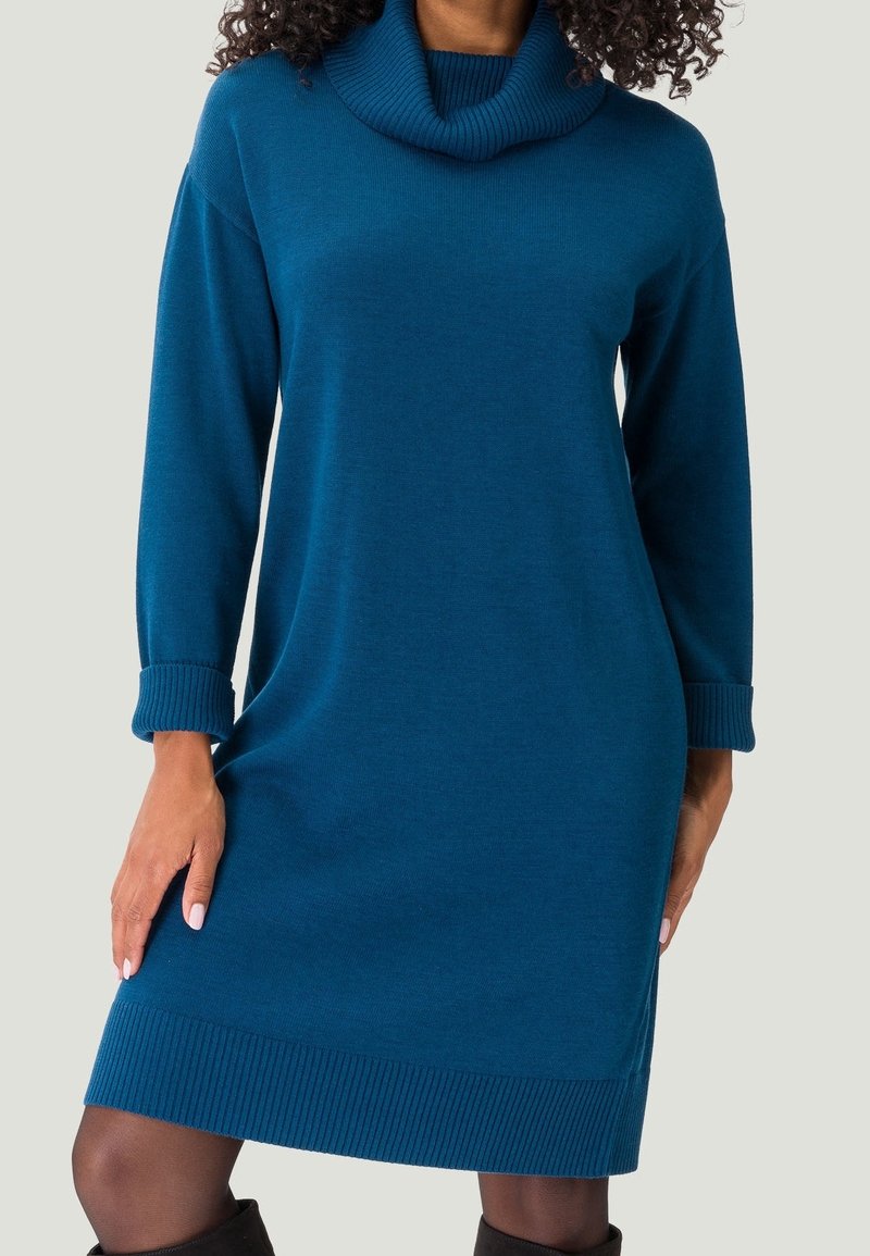 Femme portant une robe pull en maille bleu sarcelle à manches longues avec un col roulé plié et des poignets et un ourlet côtelés, et des bottes noires.