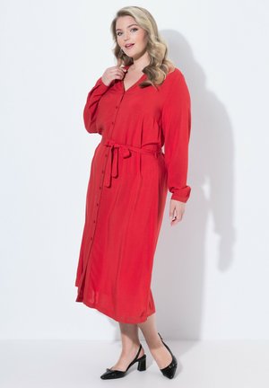 Frau mit langen blonden Haaren, die ein rotes, knöpfbares Midikleid mit Taillengürtel und schwarzen flachen Slingback-Schuhen trägt und steht.