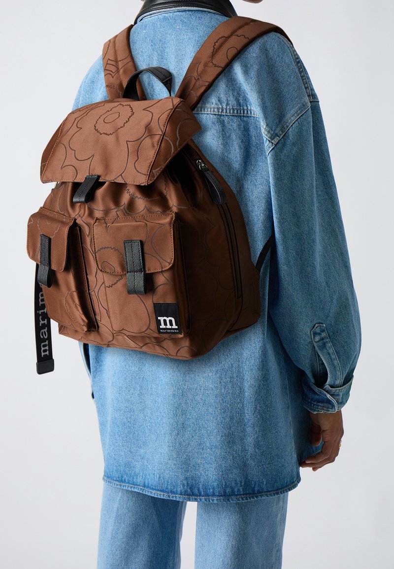 Personne portant un sac à dos marron à motifs floraux avec des poches avant et des fermetures noires, par-dessus une veste en jean bleu clair et un jean.