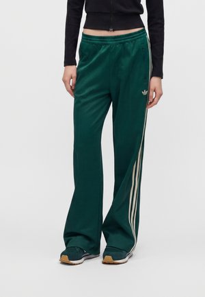Persoană purtând pantaloni de trening Adidas verde închis cu dungi albe pe părți, top negru și adidași verde închis cu tălpi bej.