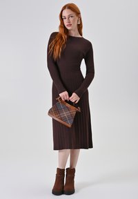 Abito lungo a maniche lunghe di colore marrone a coste, fino al ginocchio. Gli accessori includono una borsa a quadri con accenti marroni e stivaletti alla caviglia in suede marrone.