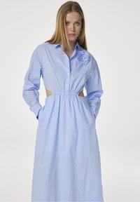 Lichtblauwe gestreepte shirtjurk met lange mouwen, een getailleerde taille, zijuitsnijdingen en een decoratieve bloemenaccent op de schouder.