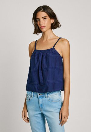 Top sin mangas azul marino con tirantes trenzados, escote fruncido y detalles de costura horizontal; combinado con jeans de denim azul claro.