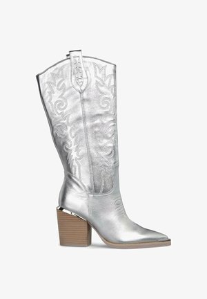 Bota vaquera metálica plateada con puntera afilada y patrones florales en relieve, que cuenta con un tacón cuadrado y costuras decorativas en el cañón.