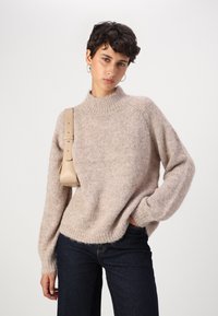 Beige knitted sweater with a high neckline, ribbed hem, and long sleeves. Paired with dark jeans and a small beige shoulder bag.