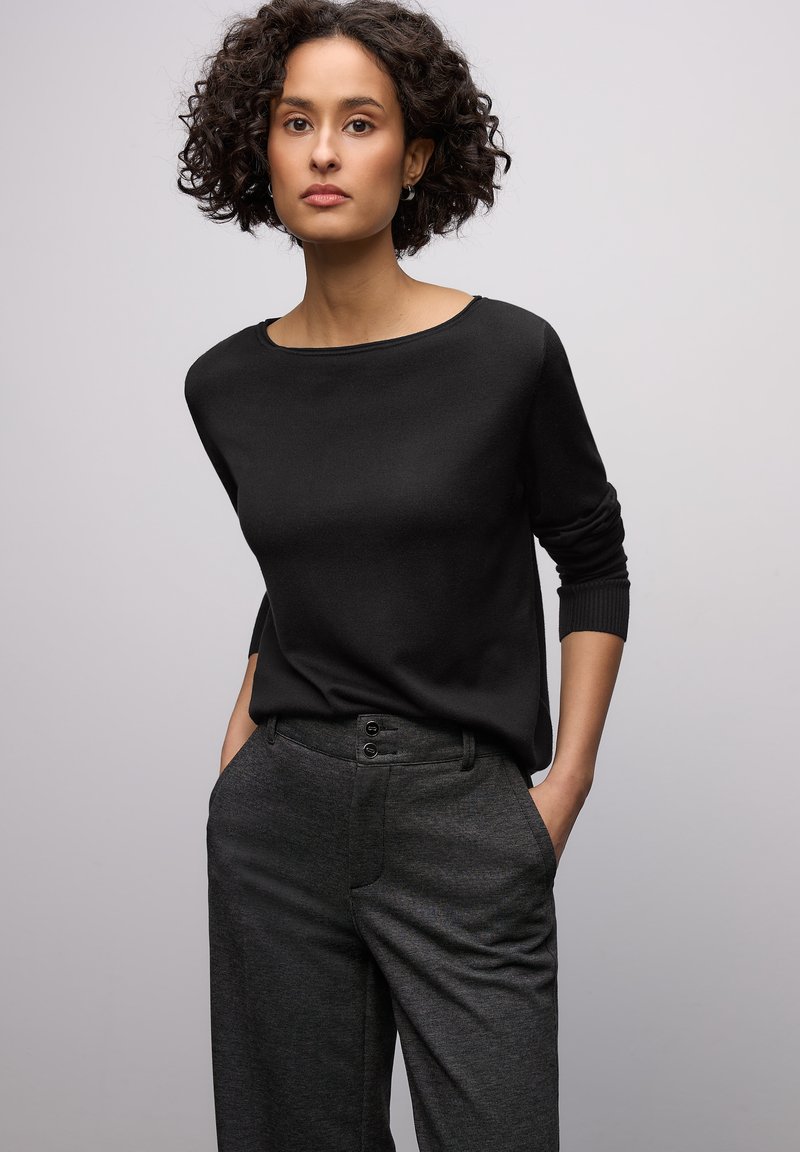 Schwarzer Pullover mit langen Ärmeln und Bootsausschnitt, kombiniert mit grauen Hosen mit feiner Textur und doppelter Knopfverschluss an der Taille.