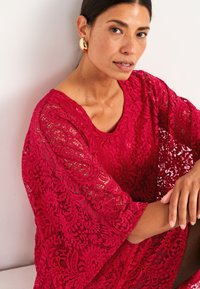 Röd spetsblus med vida ärmar, med intrikata blommönster. Tyget är texturerat och semi-transparent, vilket framhäver designdetaljerna.