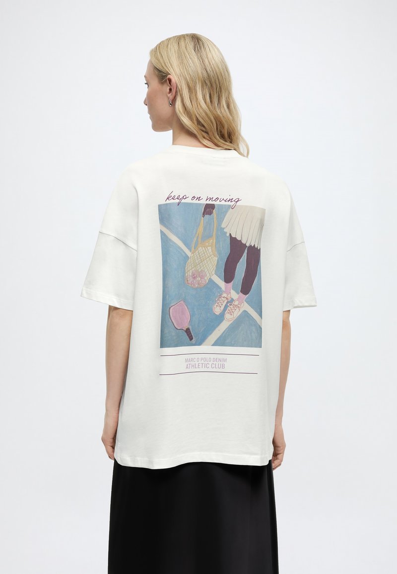 Mujer con cabello rubio, vistiendo una camisa blanca de gran tamaño con una ilustración y texto de temática de tenis que dice "keep on moving" en la parte posterior.