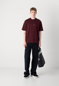Burgundy cotton t-shirt with short sleeves featuring "Calvin Klein" logo, paired with dark denim trousers and a black tote bag.