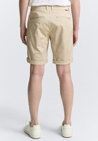 Beige Baumwollshorts mit umgeschlagenen Säumen, zwei Gesäßtaschen und einer glatten Textur. Getragen mit weißen Sneakern vor einem hellgrauen Hintergrund.