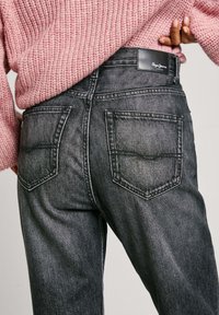 Jeans de mezclilla negra con corte recto. Presenta dos bolsillos traseros, un ligero desgaste y una etiqueta de cuero en la cintura con el nombre de la marca.
