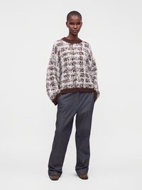 Personne portant un pull marron et blanc à motifs, un pantalon ample gris foncé et des chaussures marron, debout devant un fond blanc uni.