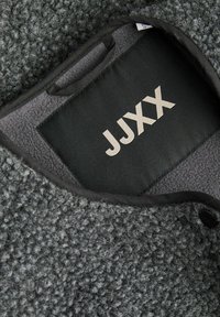 Sivý fleecový odev s textúrovaným povrchom. Obsahuje čierny textilný štítok s nápisom "JJXX" v svetlosivej farbe. Zahŕňa malú textilnú slučku.