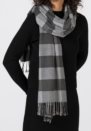 Femme portant un haut noir à manches longues et une écharpe grise à carreaux avec franges, enroulée lâchement autour du cou, sur un fond clair uni.