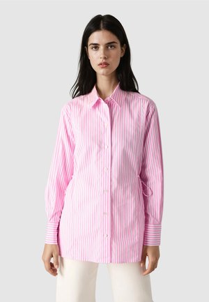 Woman wearing a long-sleeve pink and white vertical striped button-up shirt with a collar, standing against a plain light background.