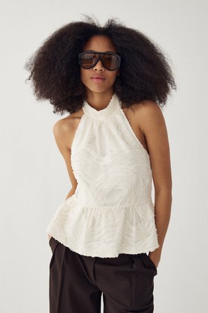 Vrouw met grote krullende haren draagt oversized donkere zonnebril, mouwloze witte getextureerde haltertop en donkere broek, handen in haar zakken.