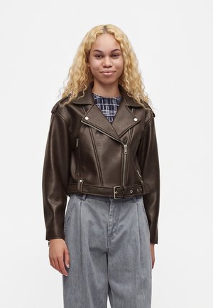 Jeune personne aux longs cheveux blonds bouclés, portant une veste en cuir marron foncé, une chemise à carreaux et un pantalon gris ample, debout devant un fond blanc.