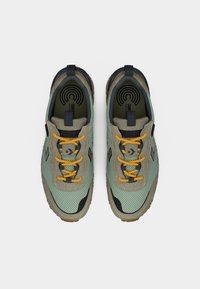 Ζευγάρι πράσινα και μπεζ αθλητικά παπούτσια με κίτρινες κορδόνια, μαύρες λεπτομέρειες και εσωτερικές σόλες με λογότυπο, απεικονιζόμενα από πάνω σε λευκό φόντο.