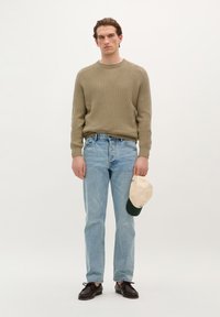 Beige knitted sweater, light blue jeans, black lace-up shoes, and a cream and green cap held in hand. Minimalist design with textured fabric.