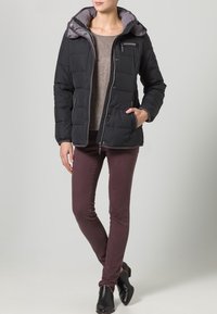 Černá zateplená bunda s šedou kapucí, zapínání na zip, boční kapsy a prošívaná textura, oblečená přes hnědý svetr a vínové kalhoty.