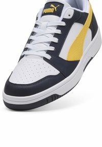 Puma sneakers i en navy og hvid farveskema, med en stor gul stribe, perforeret tåboks og hvide snørebånd. Gummisål.