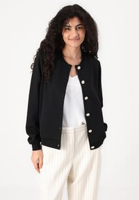 Black textured bomber jacket with silver buttons and a round neckline, worn over a white top and paired with cream striped trousers.