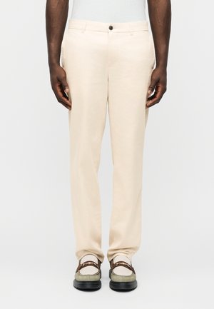 Άνδρας που φοράει ανοιχτό μπεζ παντελόνι με ίσια γραμμή και πράσινο-γκρι μοκασίνια με καφέ λουριά, στέκεται μπροστά σε απλό φόντο.