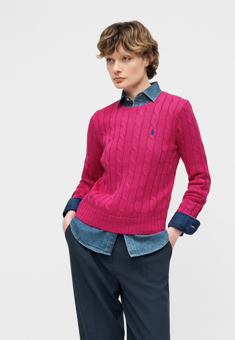 Femme aux cheveux courts portant un pull en tricot torsadé rose vif par-dessus une chemise en denim avec un pantalon foncé, debout les mains dans les poches.