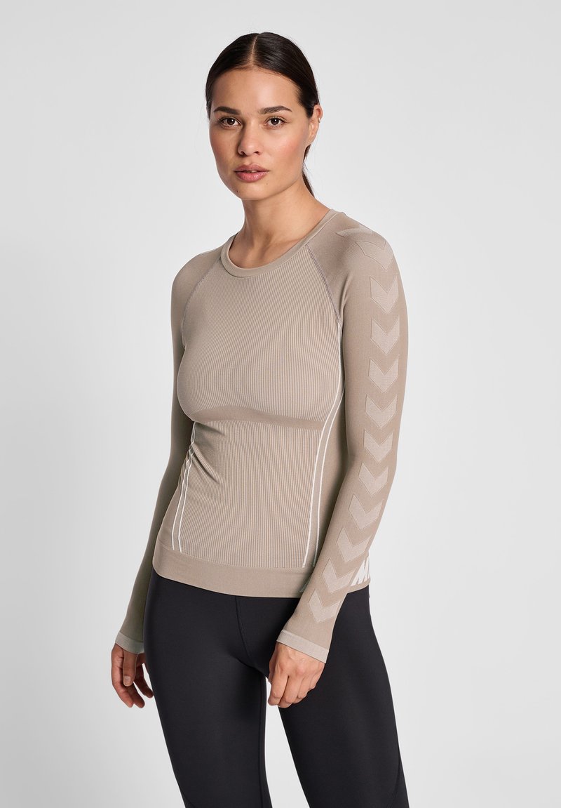 Femme portant un haut beige cintré à manches longues avec des motifs en chevrons sur les manches et un legging noir, debout devant un fond clair uni.