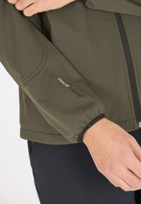 Lähikuva henkilön kädestä, joka pitää päällään oliivinvihreää takkia, jossa hihellä on teksti "Windpro 8000" ja musta vetoketju.