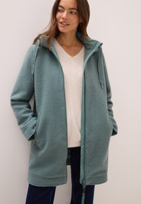 Vihreä fleece-takki, jossa on vetoketjukiinnitys, säädettävä huppu, sivutaskut ja pehmeä pinta, yhdistettynä vaaleanbeigeen paitaan ja tummiin farkkuihin.