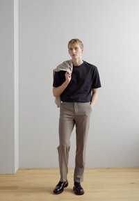 Tricou negru cu mânecă scurtă, pantaloni eleganți bej, pulover gri aruncat peste umăr și pantofi negri; design minimalist și culori neutre.