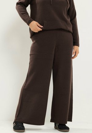Person trägt dunkelbraune, weit geschnittene Strickhose mit passendem Strickoberteil und schwarzen Sneakern, steht vor einem schlichten Hintergrund.